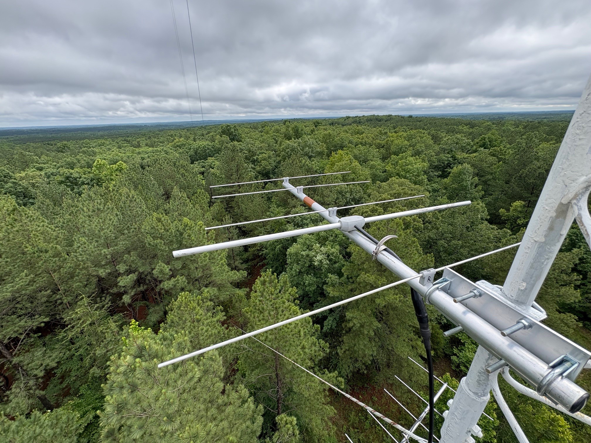 View above trees under white clouds with parts of a tall tower and antennae visible.